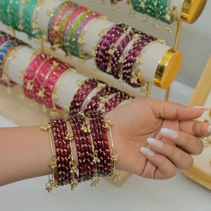 Puede incluir: Una exhibición de pulseras coloridas en varios tonos, incluyendo rojo, rosa, verde y morado. Las pulseras están adornadas con pequeños detalles dorados y se presentan en un expositor blanco. Una mano que lleva un conjunto de pulseras rojas con detalles dorados está en primer plano.
