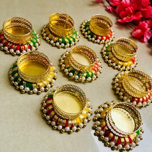 Pode incluir: Nove castiçais decorativos com bordas e bases de metal dourado. As bases são adornadas com fio vermelho, verde e amarelo, acentuadas com contas brancas peroladas. Os castiçais estão dispostos sobre uma superfície clara.