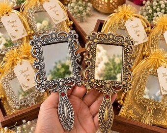 May include: A silver-toned and a gold-toned handheld mirror, each featuring an ornate, embossed frame with a small red accent at the base of the handle. Each mirror is packaged in a gold organza drawstring bag with a tag that reads Thank You.