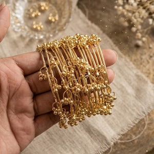 May include: A collection of gold-coloured bangles with a detailed design. Each bangle has small, round decorations and is held in a hand. The jewellery is presented on a neutral-toned fabric, with more gold-coloured jewellery visible in the background.