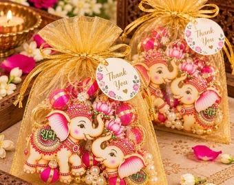 May include: Two gold organza drawstring favor bags filled with decorative elephant-shaped ornaments, pink spheres, and white beads. Each bag features a round white tag with floral accents and the text Thank You in gold script.