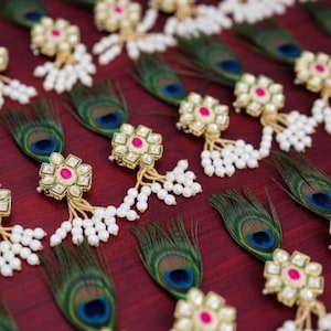May include: Arrangement of decorative items featuring peacock feathers and floral-shaped embellishments. Each piece has a gold-colored base with a pink center and white pearl accents. The feathers are green with a blue and black eye.