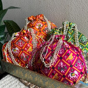 May include: A collection of colorful drawstring bags with intricate geometric patterns. The bags are in shades of red, orange, pink, and green, adorned with pearl-like beaded straps. They are displayed on a decorative tray.