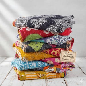 May include: A stack of colorful handmade Kantha quilts. The quilts feature various patterns and colors, including red, blue, yellow, and black. A tag reads "Handmade Kantha Quilts Unique & Eco-Friendly."