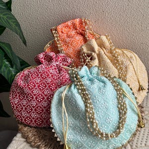May include: Four embroidered drawstring bags in red, orange, light blue, and cream. Each bag features intricate white floral patterns and is adorned with beaded accents and gold trim. The bags are displayed on a woven surface.