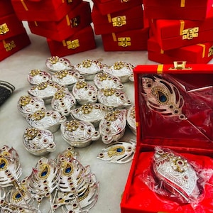 May include: An assortment of ornate silver and gold-toned jewelry pieces, including peacock feather designs and small, round containers. Many items are displayed in red velvet boxes with gold clasps, suggesting a theme of luxury and gifting.