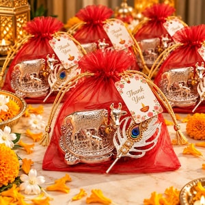 May include: A red organza drawstring gift bag containing a silver-toned cow and calf figurine on a circular base and a silver-toned peacock feather ornament. A white tag attached to the bag features the text Thank You and a small diya icon.