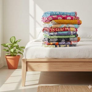 May include: A stack of colorful, patterned quilts on a bed with a light wood frame. The quilts display a variety of colors, including blue, pink, yellow, and green. A potted plant sits beside the bed.