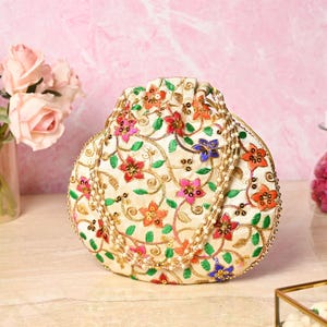 May include: An off-white, embroidered pouch with a drawstring closure. The pouch is adorned with colorful floral patterns in red, orange, pink, and blue, with gold-colored accents. The bag is likely used for carrying small items.