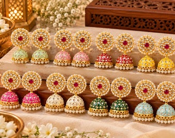 May include: A collection of sixteen jhumka earrings arranged in two rows. Each earring features a circular stud with a central red stone surrounded by white beads, connected to a bell-shaped drop in various colors including green, pink, red, yellow, white, and blue, all accented with gold-tone metal and small white pearls.