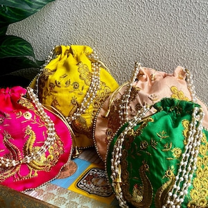 Könnte beinhalten: Vier farbenfrohe Kordelzugbeutel in Pink, Gelb, Pfirsich und Grün. Jede Tasche hat goldene Stickereien eines Pfaus und Blumenmuster. Weiße Perlenschnüre dienen als Kordelzüge und Akzente. Die Taschen sind auf einem dekorativen Tablett angeordnet.