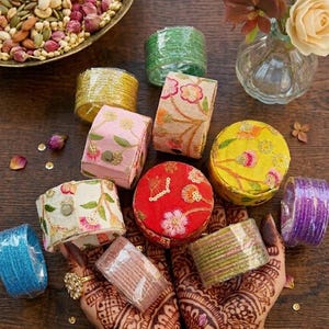 May include: An assortment of colorful, decorative boxes and bangles. The boxes are round and rectangular, adorned with floral embroidery and sequins in shades of pink, yellow, and red. The bangles are wrapped in cellophane and come in various colors, including blue, green, and purple.