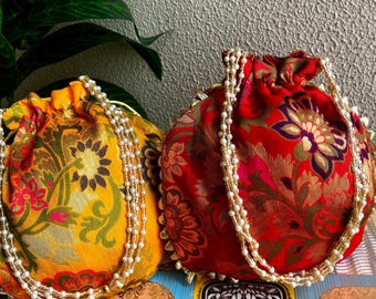 Könnte beinhalten: Zwei dekorative Beutel mit Kordelzug. Einer ist gelb mit einem Blumenmuster, der andere rot mit einem ähnlichen Design. Beide haben perlenartige, perlenbesetzte Griffe und Verzierungen. Die Beutel befinden sich auf einer gemusterten Oberfläche.