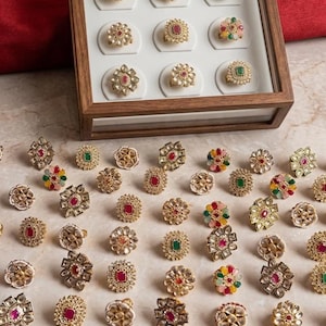 May include: A collection of gold-toned rings with various gemstone settings, including red, green, and multi-colored stones. Some rings are displayed in a wooden box, while others are scattered on a marble surface. The rings feature intricate floral and geometric designs.