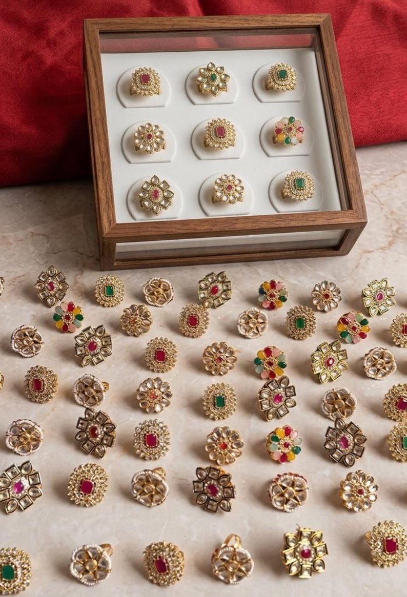 May include: A collection of gold-toned rings with various gemstone settings, including red, green, and multi-colored stones. Some rings are displayed in a wooden box, while others are scattered on a marble surface. The rings feature intricate floral and geometric designs.