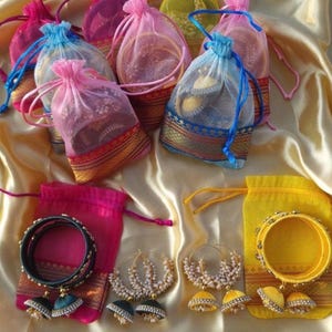 May include: An assortment of colorful jewelry, including bangles, hoop earrings, and jhumka earrings, displayed in sheer drawstring bags. The bags are in various colors like pink, blue, and yellow, with gold trim. The jewelry is set on a gold satin surface.
