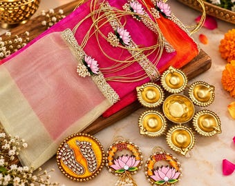May include: A vibrant pink and orange fabric pouch with gold metallic trim and lotus flower accents, accompanied by a gold-toned seven-wick oil lamp and three circular decorative ornaments featuring lotus flowers and footprints.