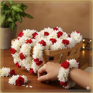 May include: A basket of white and red floral garlands. The garlands are made of white, fluffy flowers with red rose accents. A hand wears a matching wrist garland. The garlands are displayed on a wooden surface. The theme is floral.