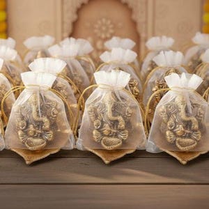 Peut inclure: Une collection de petites statues de Ganesha dorées, chacune présentée dans un sac en tulle blanc. Les statues reposent sur des socles décoratifs dorés, créant une présentation élégante et spirituelle. L'arrière-plan présente un design architectural orné flou.