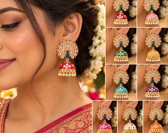 May include: A collection of jhumka earrings featuring a gold-tone peacock-shaped stud with clear crystals and a central blue stone. The bell-shaped drop is decorated with colorful enamel and small white beads along the bottom edge.