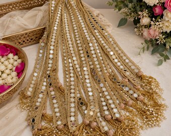 May include: A long, decorative gold ribbon trim featuring a repeating pattern of small, circular mirrors embedded within a woven metallic fabric. The ends of the trim are finished with gold tassels, small gold beads, and dangling white pearls.