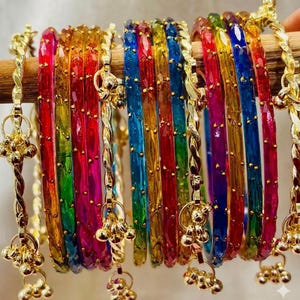 May include: A collection of colorful glass bangles in a rainbow of hues, including red, orange, yellow, green, blue, and purple. Each bangle is adorned with gold-colored metal accents and dangling charms, creating a vibrant and eye-catching display.