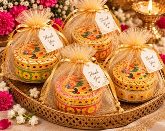 May include: Four round metal tins with colorful peacock and floral patterns, each enclosed in a gold organza drawstring gift bag with a white tag printed with the text Thank You and a small heart icon, arranged on a gold metal tray.