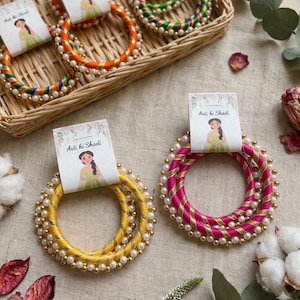 以下が含まれることがあります： パールアクセントが施されたカラフルなバングル。バングルは黄色、ピンク、オレンジ、緑の色合いです。各バングルには「Arti ki Shadi」の文字と女性のイラストが描かれたタグが付いています。