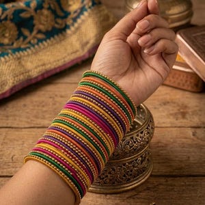 May include: A stack of colorful bangles in shades of green, orange, pink, purple, yellow, and gold. The bangles are worn on a wrist, with a wooden surface in the background. The bangles are adorned with small gold beads.