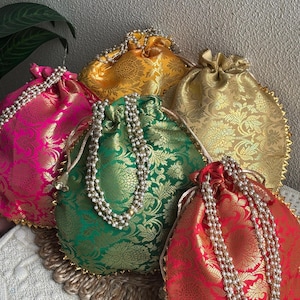 Puede incluir: Cinco coloridos bolsos con cordón en fucsia, dorado, verde, rojo y amarillo. Cada bolso presenta un diseño floral dorado, un cordón de cuentas y un ribete dorado. Fabricados con tela estampada.