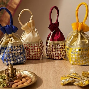 May include: Four decorative drawstring pouches in blue, cream, burgundy, and yellow. Each pouch has a gold base, embellished with mirrored accents. The pouches are arranged on a wooden surface, with a brass plate holding nuts and a small statue.