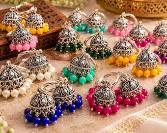 May include: A collection of silver-toned jhumka earrings featuring intricate metalwork domes and dangling beads in various colors including yellow, pink, white, green, blue, and magenta, arranged on a textured surface.