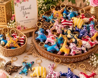 May include: A collection of plush animal keychains featuring various patterns including leopard print, floral, and stripes. The keychains are displayed in a wooden tray and a small woven basket, accompanied by a sign that reads Thank You FOR CELEBRATING WITH US.