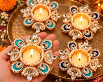 May include: Four circular candle holders featuring a central lit tea light surrounded by a ring of small white beads and petal-shaped clear crystals. The holders have alternating teal and dark blue fabric accents arranged in a floral pattern.