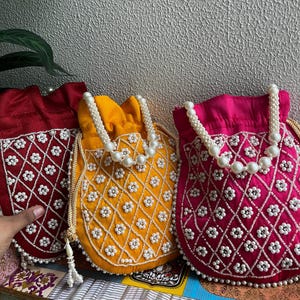 Puede incluir: Tres bolsos con cordón y cuentas en rojo, amarillo y rosa. Cada bolso presenta un patrón floral con cuentas blancas y un asa con cuentas de perlas. Los bolsos están hechos de una tela suave y texturizada, ideales para objetos pequeños.