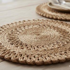 May include: Two round, woven jute placemats. The placemats are a natural tan color with a spiral design in the center and a scalloped edge. One placemat is in the foreground, and another is in the background with a plate and silverware.