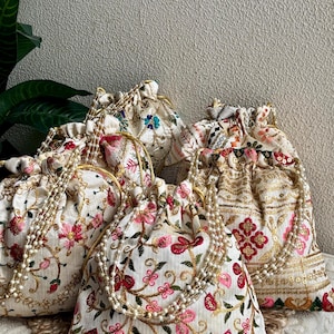 May include: A collection of embroidered drawstring bags. The bags feature floral patterns in shades of pink, red, and gold on a cream background. Each bag has a beaded strap and gold trim.