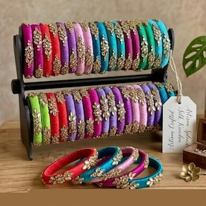 May include: A display of colorful silk bangles with gold-toned floral embellishments. The bangles are arranged on a black stand, showcasing a variety of colors including pink, red, purple, and teal. Some bangles are displayed separately. The image has a warm, inviting aesthetic.