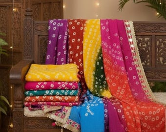 May include: A stack of colorful, patterned scarves in shades of yellow, pink, orange, green, and blue rests on a wooden bench. The scarves have a dotted design and are trimmed with gold and tassels. A wooden screen is in the background.