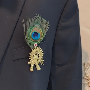 Puede incluir: Un broche dorado con una deidad tocando la flauta, adornado con una pluma de pavo real y una peque&ntilde;a gema roja. El broche est&aacute; sujeto a una blazer azul marino oscuro, creando un contraste llamativo.