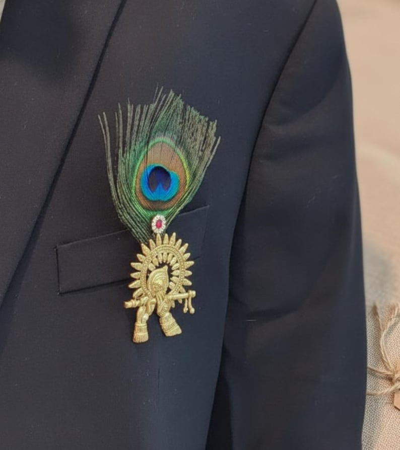 Puede incluir: Un broche dorado con una deidad tocando la flauta, adornado con una pluma de pavo real y una peque&ntilde;a gema roja. El broche est&aacute; sujeto a una blazer azul marino oscuro, creando un contraste llamativo.