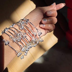 May include: A collection of silver-toned bracelets, each featuring a series of small, decorative elements. The bracelets are displayed on a person's arm, showcasing their design and style. The bracelets have a delicate, ornate appearance.