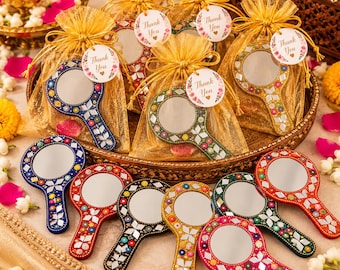 May include: A collection of round handheld mirrors featuring colorful floral patterns and small reflective mirror tiles on the handles and frames. Several mirrors are packaged in gold organza drawstring bags with round Thank You tags.