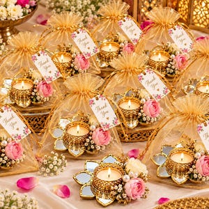May include: A collection of gold-toned metal lotus-shaped candle holders with mirrored petals, each holding a lit tea light, adorned with pink roses and white baby's breath, packaged in gold organza bags with Thank You tags.