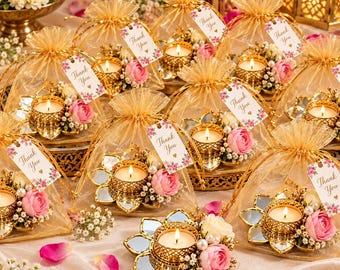 May include: A collection of gold-toned metal lotus-shaped candle holders with mirrored petals, each holding a lit tea light, adorned with pink roses and white baby's breath, packaged in gold organza bags with Thank You tags.