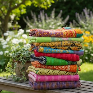 May include: A stack of colorful, folded quilts rests on a wooden bench. The quilts display a variety of patterns and colors, including red, green, yellow, and pink. The background features lush greenery and flowers.
