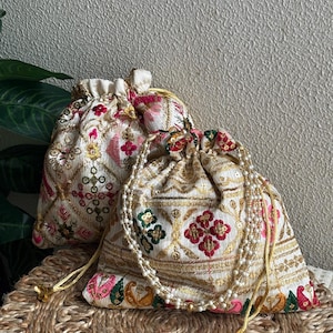 Op de afbeelding: Twee crèmekleurige trekkoordtassen met ingewikkelde borduursels in goud, roze, rood en groen. De tassen hebben een bloemenpatroon en zijn versierd met een parelkoorden trekkoord. Ze liggen op een geweven mat.