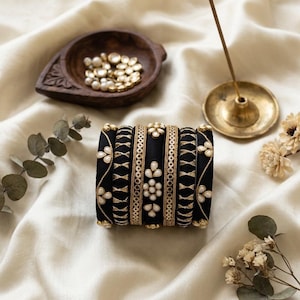 May include: A set of black bangles with gold and white floral embroidery. The bangles are arranged on a cream-colored fabric, alongside a wooden bowl with gold-colored beads, a brass incense holder, and dried flowers.