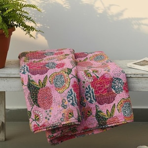 May include: Pink floral quilt with a vibrant pattern of flowers and leaves. The quilt is folded on a white bench next to a potted plant and a book. The quilt's design includes red, yellow, and blue accents.