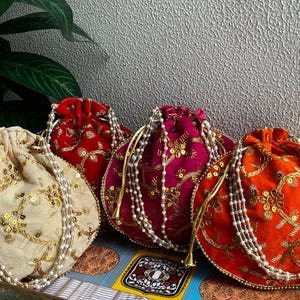 May include: Four velvet drawstring pouches in cream, red, fuchsia, and orange. Each pouch is decorated with gold embroidery and sequins, and has a pearl beaded drawstring. The pouches are displayed on a decorative box.